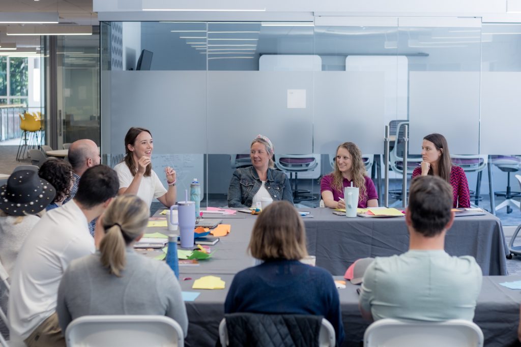 A group of deans of students engaged in a discussion around a table in a modern office setting at the deans roundtable annual conference, deans roundtable membership