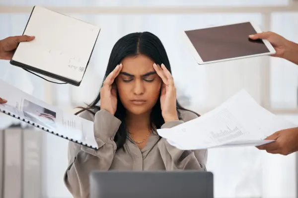 An educator at a desk, clearly frazzled, as they are being handed 4 different things to work on, embodying educator burnout